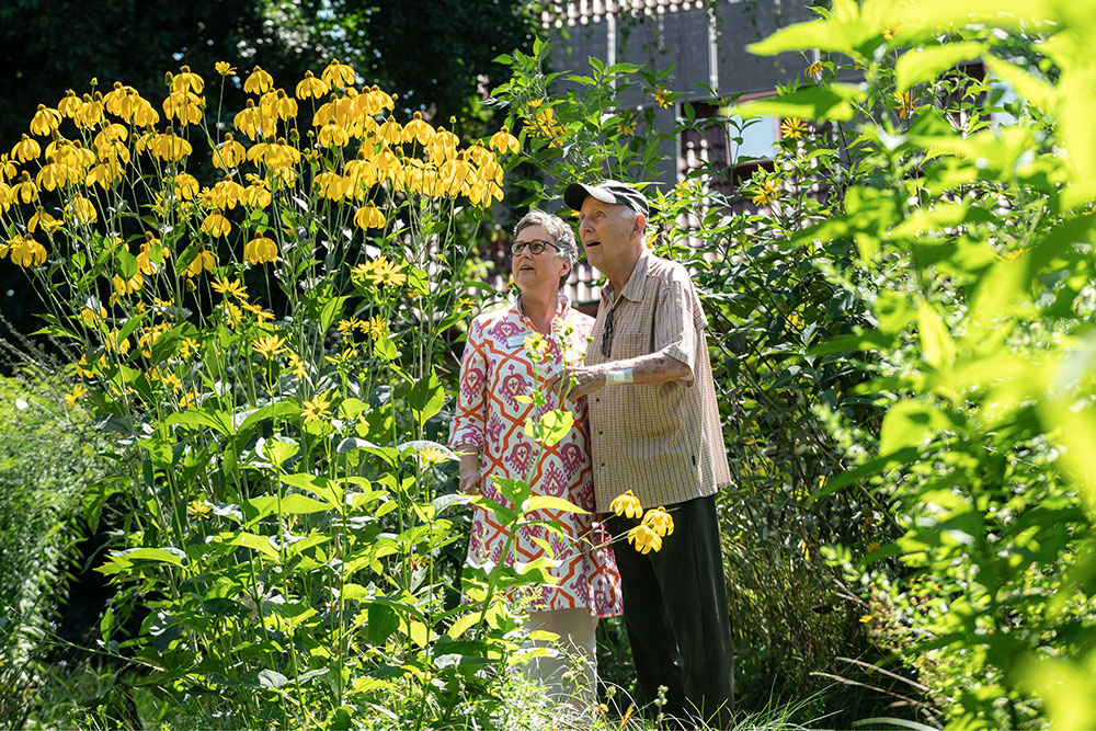 Eine ältere Dame und ein älterer Herr, die zwischen Blumenstauden im Garten des Ricam Hospizes, Orchideenweg in Berlin stehen.